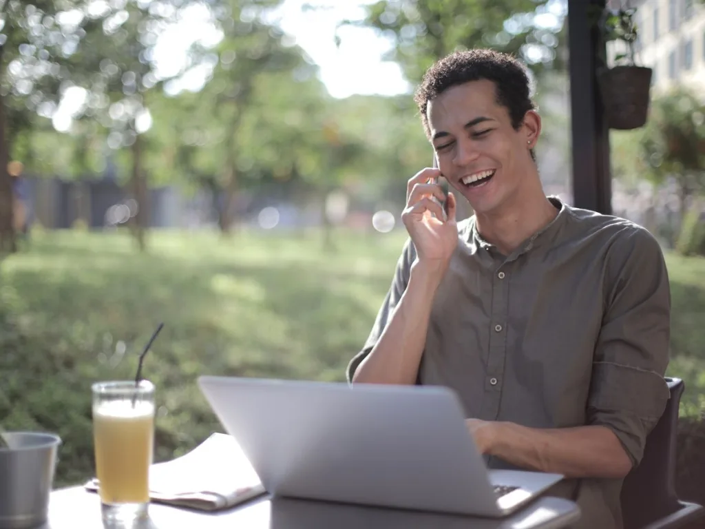 Jonge man zit aan een tafel met een laptop te werken en zit aan de telefoon