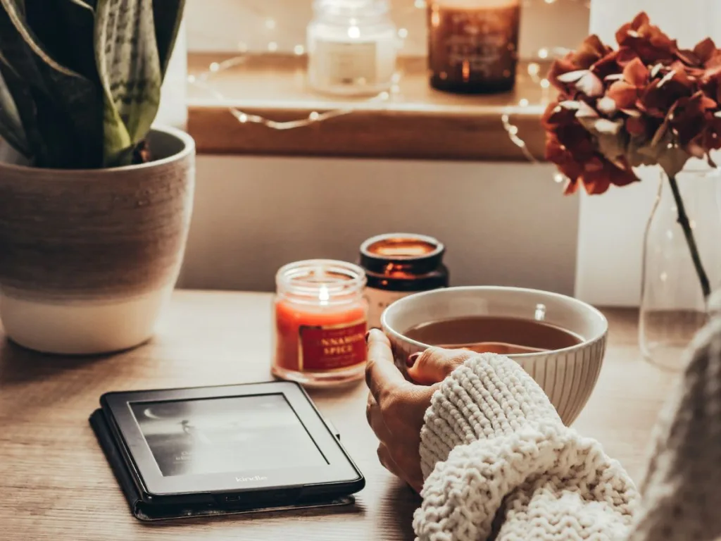 Vrouw zit in een knus hoekje in huis met een kop chocolademelk aan een tafel met een e-reader en kaarsen en kerstverlichting om haar heen