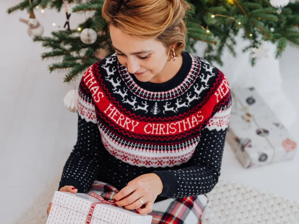 Vrouw met foute kersttrui aan zit voor de kerstboom een cadeau uit te pakken