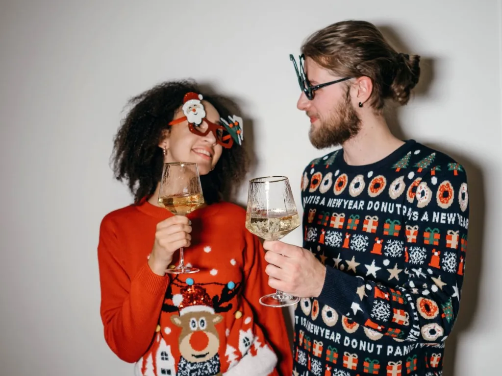 Man en vrouw proosten met een glas wijn en hebben beiden een foute kersttrui aan